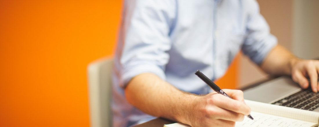 Person writing notes while looking at a laptop