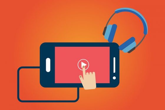 A hand clicks play on a video on a smartphone; headphones rest beside it.