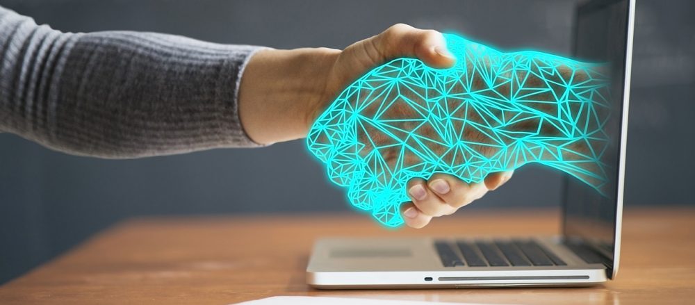 A wireframe hand extends from a laptop screen and shakes a human hand.